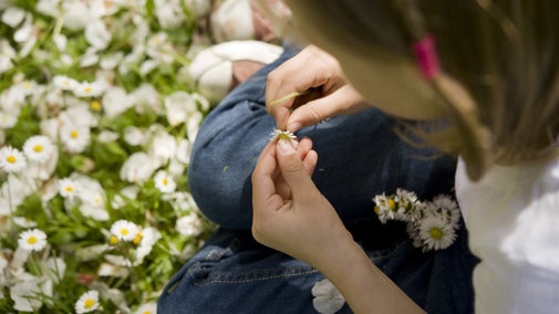 Close-up of a child sitting in a grassy meadow filled with white daisies, carefully threading flowers together to make a daisy chain on a sunny day.